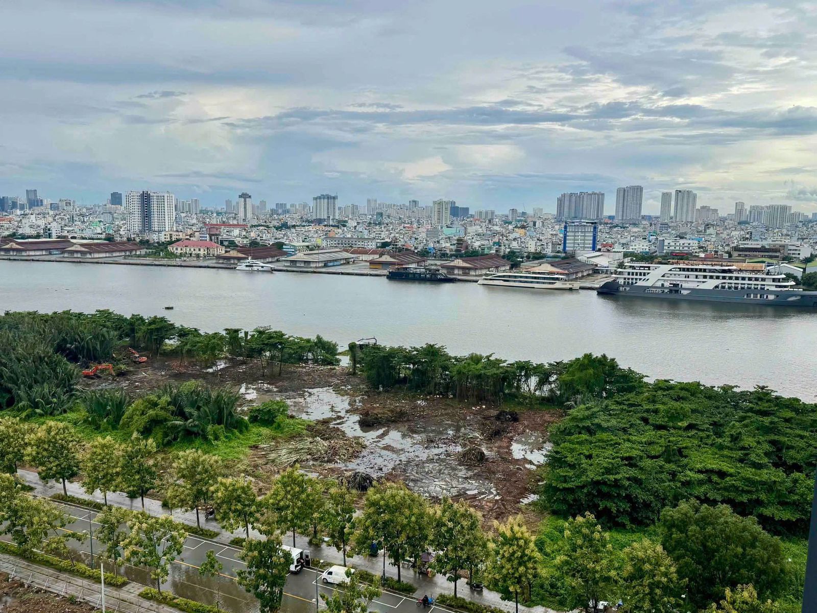 Ảnh chụp từ trên cao một dải đất ven sông: một khoảng rộng vừa bị phát quang còn bùn và vũng nước, hai máy xúc màu cam ở mép trái, phía trước là con đường có hàng cây; bên kia sông là dãy nhà kho mái thấp và mấy tàu du lịch neo sát bờ.